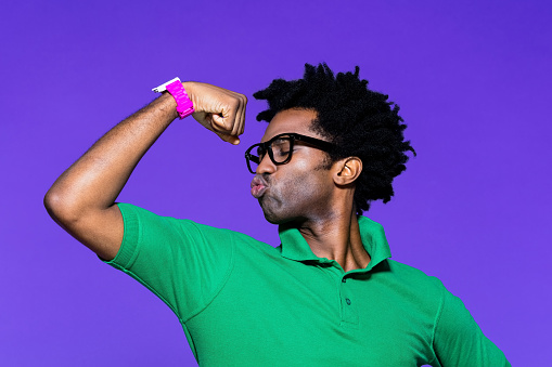 Portrait of confident afro american young man wearing green polo shirt and nerdy glasses, raising arms and flexing muscles. Studio shot on violet background.