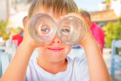 Cute boy looking at glasses as of binoculars. Kid having fun outside. Smiling child waiting for order at cafe outdoor. Happy childhood concept. Looking through binoculars. Smart quarantine