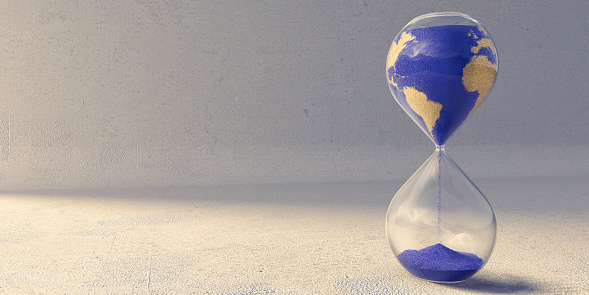A close up image of an hourglass filled with blue and yellow sand, in the pattern of the countries of the earth. Sand is slowly trickling from the top to pile up in the bottom chamber over time. The timer sits on a rough textured concrete floor near wall.