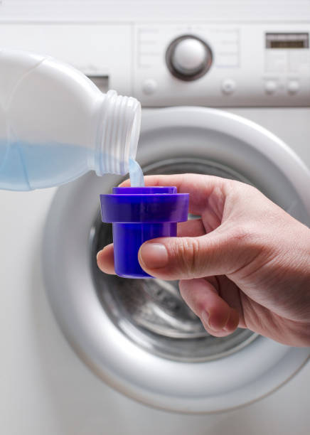 vertical image.man holding a cup and pouring liquid laundry detergent against washing machine.household routine - liquid soap blue plastic textile imagens e fotografias de stock
