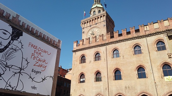 Bologna, Italy - May 26, 2020: Phase 2 Covid-19 day after. Clock tower of Palazzo d'Accursio or Comunale palace overlooking Piazza Maggiore, square. Poster for release of Patrick Zaki activist.