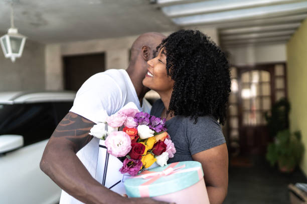husband surprising his wife with a flowers and present at home - men african descent giving flower imagens e fotografias de stock