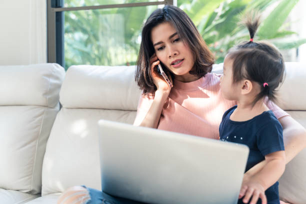 istock Asian young single mother talking on mobile phone and using computer for working from home while take care little baby girl together at home. Woman busy with urgent work. Due to Covid19 virus pandemic 1227562450