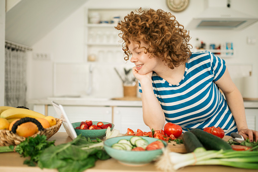 Beautiful smiling young woman with curly red hair preparing food at home in her kitchen, while watching a food channel on YouTube using her digital tablet