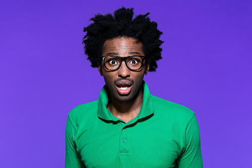 Portrait of terrified afro american young man wearing green polo shirt and nerdy glasses, staring at camera. Studio shot on violet background.