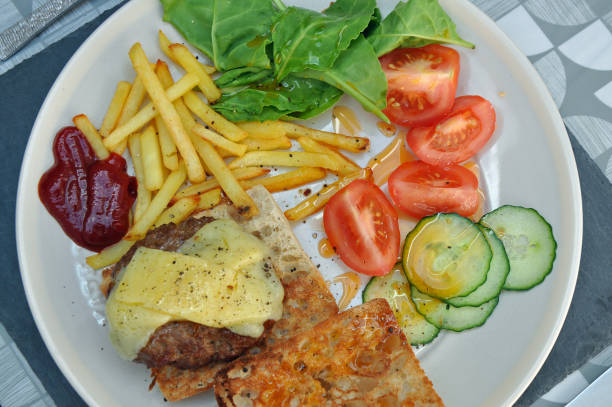 Beef burger and chips on a plate with bun cheese and salad, including spinach leaves, tomatoes, cucumber at ketchup. Authentic home cooked meal shot. Billericay, Essex, United Kingdom, May 27, 2020