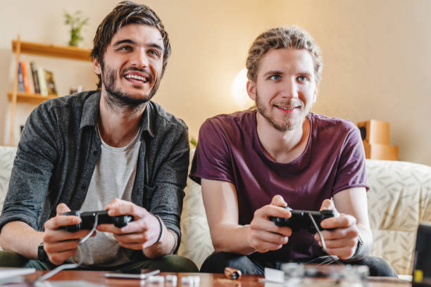 Two young smiling men playing video games while sitting on sofa at home