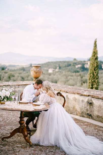 hochzeit in einer alten weinkellerei villa in der toskana, italien. das hochzeitspaar sitzt am esstisch auf dem dach einer alten villa, der bräutigam hält die braut an den händen. - puffed sleeve stock-fotos und bilder