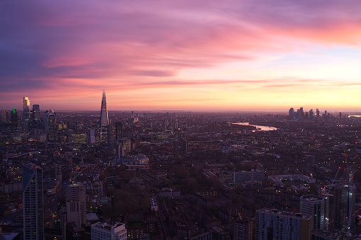 Photo shows beautiful sunrise at London where the sky is in pink and violet color. There are many houses and office building and the Thames river is reflecting the colorful sky.