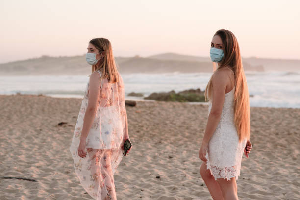 Two young women wearing protective face masks at the beach Two young women at a beautiful beach at sunset, wearing protective face masks because of COVID-19 walking beach women sadness stock pictures, royalty-free photos & images