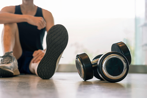 Close up pairs of black dumbbell with blurred young, fit and healthy Southeast Asian male  age 36 years sit on the floor near window with morning sunlight. Concept for healthy lifestyle, workout during stay home quarantine.