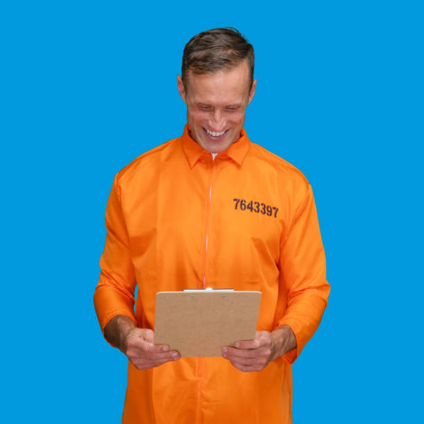 istock Criminal standing in front of blue background wearing jumpsuit and holding document 1227588510