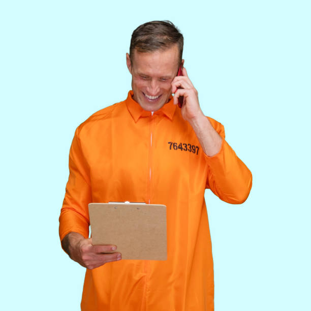 istock Criminal standing in front of blue background wearing jumpsuit and holding contract and using smart phone 1227588530