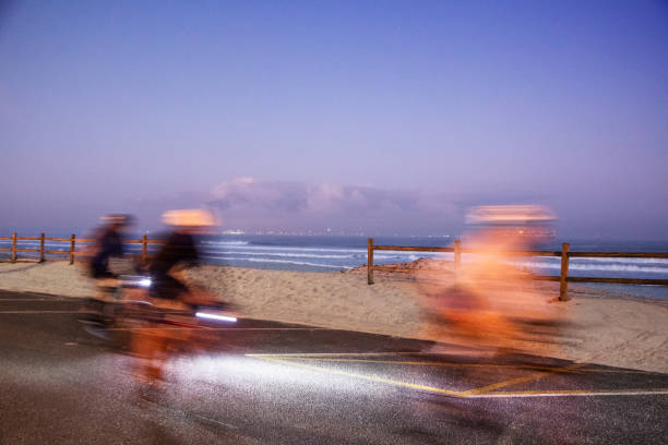 Three unrecognizable, motion-blurred people cycling with masks at dawn on Blouberg beachfront.