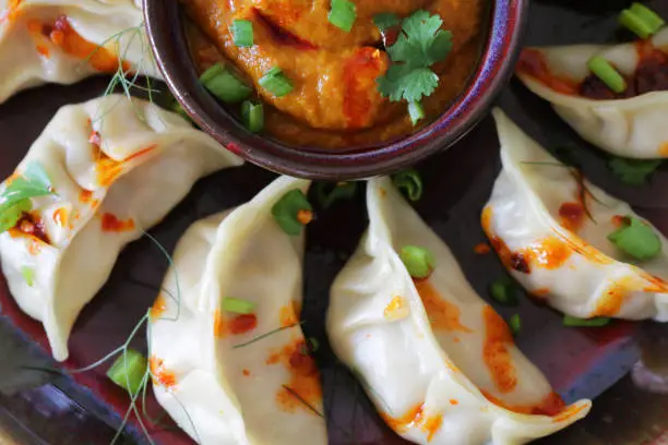 Stock photo showing an evening meal of steamed dumplings (Momos), filled with mixed vegetables and chicken drizzled with chilli oil and garnished with chopped spring onion and coriander leaves on plate with red and orange spicy dipping sauce.