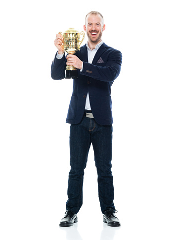 Front view of aged 30-39 years old with brown hair caucasian male manager standing in front of white background wearing jeans who is successful and winning and showing award who is in first place and holding trophy