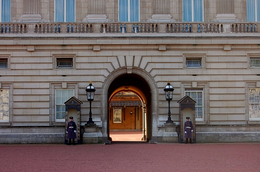 Two Buckingham Palace guards armed with rifles stand at attention in their boxes guarding an entryway into the inner courtyard of the palace