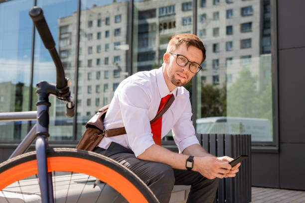 Young businessman looking at you while sitting on bench and using smartphone in urban environment after working day