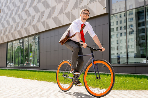 Elegant man in formalwear and eyeglasses looking forwards while going by bicycle in urban environment against exterior of business center