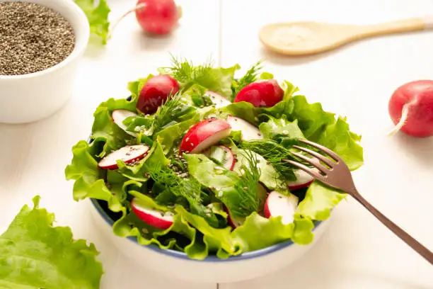 Photo of A plate with vegetarian salad stands on a wooden table. Salad of radish, lettuce, herbs and chia and sesame seeds seasoned with vegetable oil.