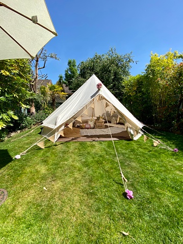 White bell tent set up to camp in private back garden on the grass during a sunny day. The tent is decorated with bunting and other decor, glamping style.