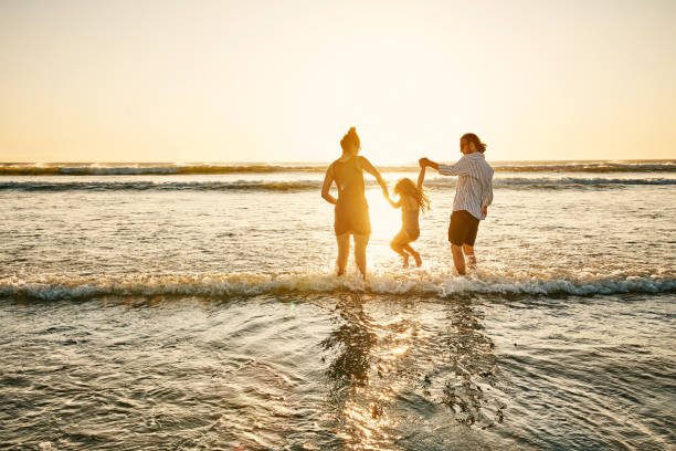 The real fun is where the waves are Shot of an adorable little girl spending the day at the beach with her parents wave jumping stock pictures, royalty-free photos & images