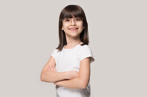 Studio portrait of confident happy brown-haired 6 years old cute girl wearing eyeglasses, standing with folded hands, isolated on grey background. Smiling smart little cutie looking at camera.