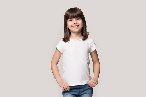 Happy six years old adorable girl in white mock up blank t-shirt on grey studio background. Portrait of smiling fringe hairstyled little cutie, holding hands in jeans pocket, looking at camera, Z generation.