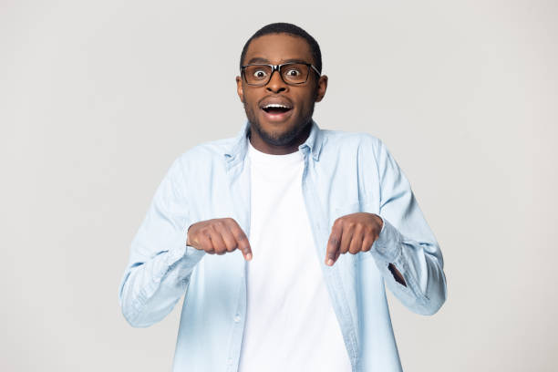 African amazed guy look at camera pointing fingers down African open mouth make big eyes gawp look at camera pointing fingers down isolated on grey background, advertising new products showing drop in prices and rebate, seasonal sales concept low stock pictures, royalty-free photos & images