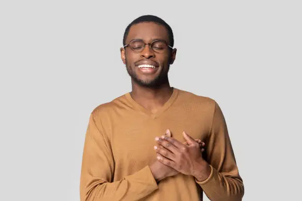 Candid handsome African guy dressed in sweater closed eyes keeps hands on chest he is cordial and friendly showing gratitude feeling thankfulness posing alone isolated on grey studio wall background
