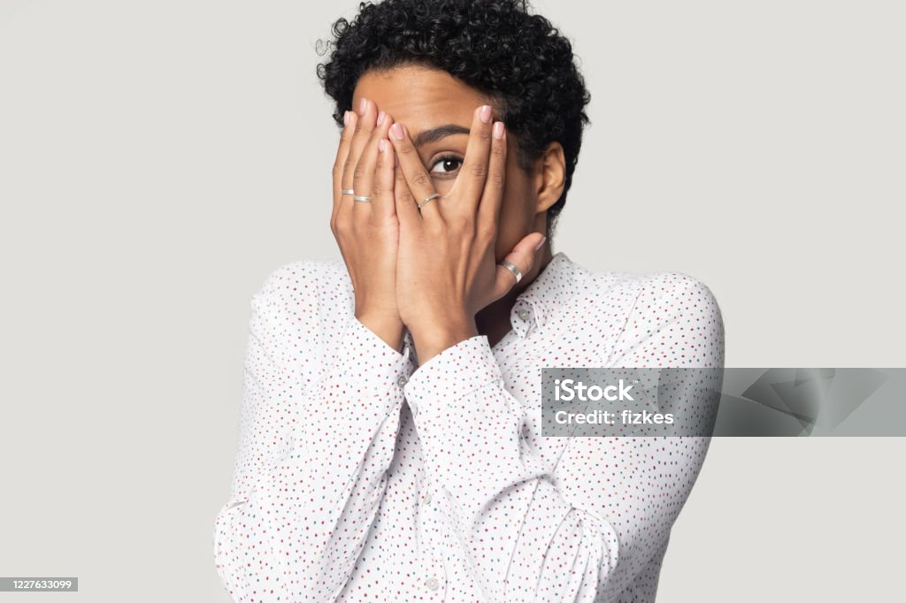 Afraid african woman hiding face with hands peeping through fingers Afraid african woman isolated on gray background looking shy or terrified covering hiding her face with hands peeping through fingers is shocked by horror movie or frightened by what she saw concept Fear Stock Photo
