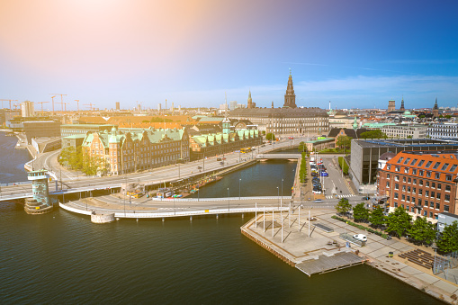 Copenhagen cityscape with the Copenhagen Stock Exchange Børsen, contemporary office buildings and residental flats interconnected to Christianshavn with bicycle bridges at the up class oceanfront, shot with drone. Dramatic cloudscape and lens flare in background.