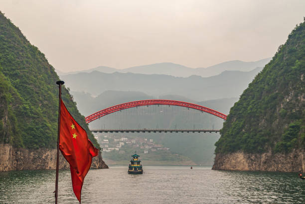 S103 bridge over Daning River with boat in Wushan, Chongqing, China. Wushan, Chongqing, China - May 7, 2010: Wu Gorge in Yangtze River. Red S103 road bow bridge over Daning River at connection with green Yangtze. Chinese flag and boat. Mountains on horizon. yangtze river stock pictures, royalty-free photos & images