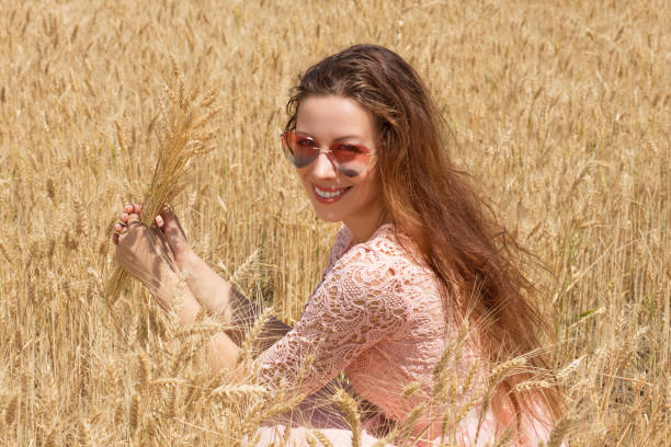 시리얼 밭에 하트 모양선글라스를 끼고 호밀 귀화환을 들고 날씨를 즐기는 밀에 앉아 웃고 있는 행복하고 아름다운 젊은 여성 - h shaped 뉴스 사진 이미지