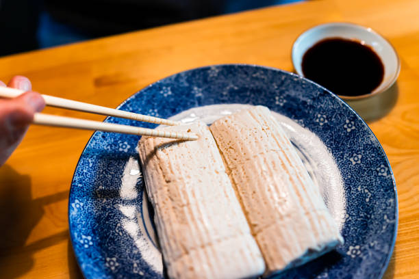 крупным планом руки проведение палочки резки японских традиционных комо тофу дофу с соевым соусом на тарелке блюдце на деревянном столе в � - chopsticks soybean japanese cuisine blue стоковые фото и изображения