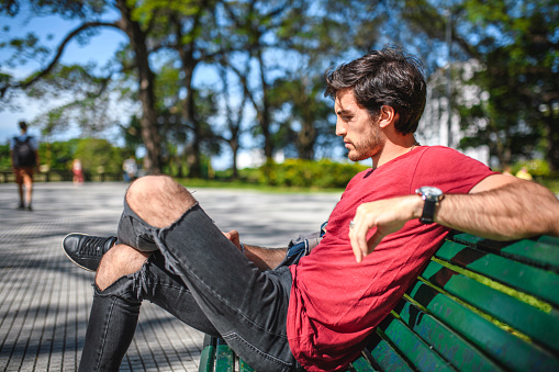 Side view close-up of Hispanic male in mid 20s relaxing on park bench and enjoying sunny Buenos Aires.