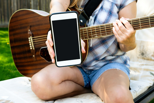 Girl playing guitar outside with mobile device in hand. Image depicts that girl is showing the viewer what app she is using to learn how to play the guitar. Screen is ready to drop in any image or branding element.