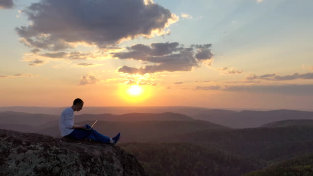 Epic drone shot of a silhouette of a young freelancer with a laptop on the edge of a rock ledge in the mountains.