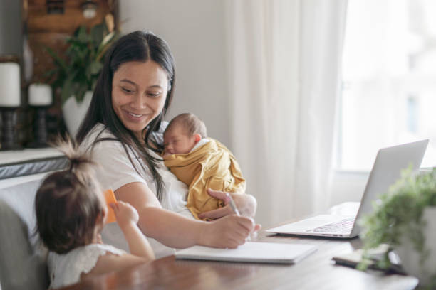 New mother holding infant trying to do work at dining room table Multi-ethnic woman sits at dining room table trying to get some work done while holding her 1 week old baby and trying to keep her toddler daughter entertained. cambodian ethnicity stock pictures, royalty-free photos & images