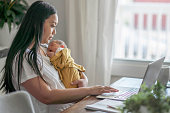 New mother holding infant trying to do work at dining room table