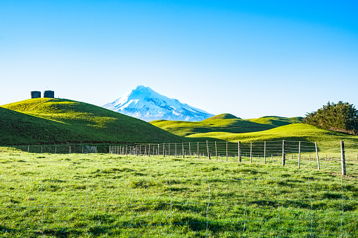 Mount Egmont snowcapped between rolling green pastures with two water tanks on hill, Taranaki New Zealand