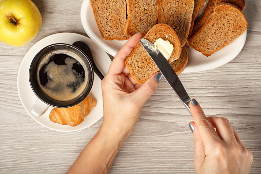 Top view of female hands spreading butter on toast at the table with cup of black coffee, croissant, bread and apple. Food and beverages for breakfast