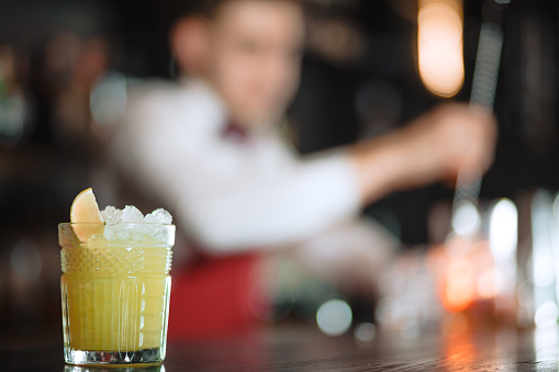 Alcohol cocktail with ice and lemon in glass isolated on blurred restaurant with barman on background.