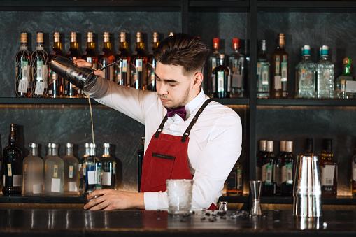 Professional bartender in uniform doing show of his work, holding two parts of metal shaker in his hands and pouring a cocktail, shelves full of bottles with alcohol on the background