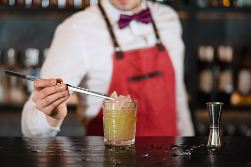 Bartender finishes decoration of his cocktail with ice cubes and lime slice using tweezers.