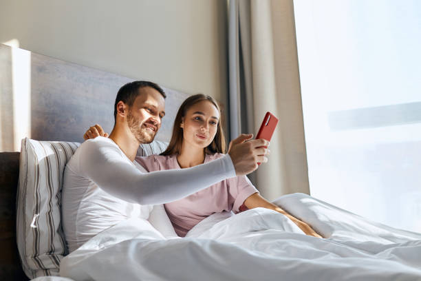 Young couple having honeymoon Cheerful emotive couple sitting in bed, stretching hand with red cell phone, looking at screen, talking to parents via skype, shot from below, family portrait mum new boyfriend stock pictures, royalty-free photos & images