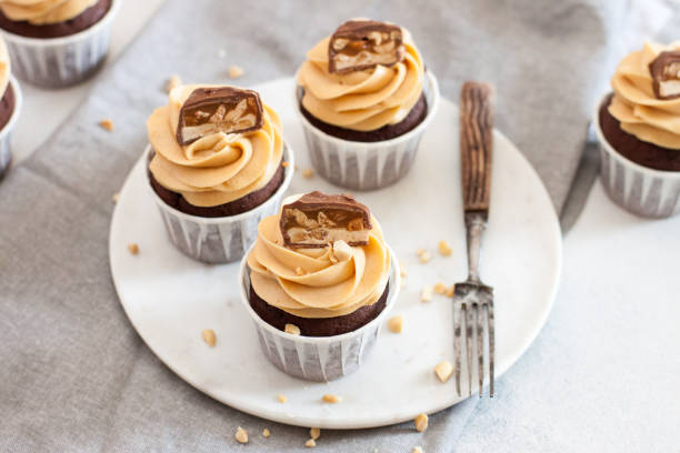 Peanut butter cupcakes with chocolate bites and chopped nuts on grey linen tablecloth.