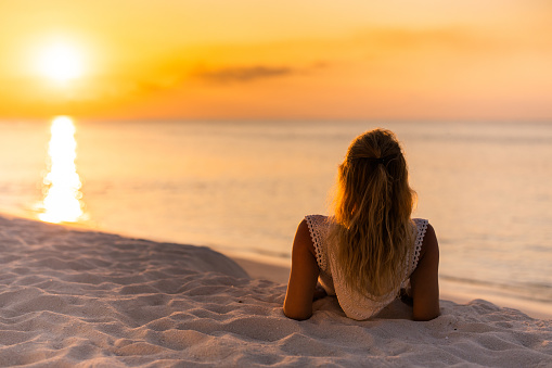Rear view of a woman relaxing in sand on the beach and looking at sea view at sunset. Copy space.