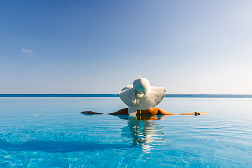 Back view of a woman with sun hat spending summer day in an infinity pool. Copy space.