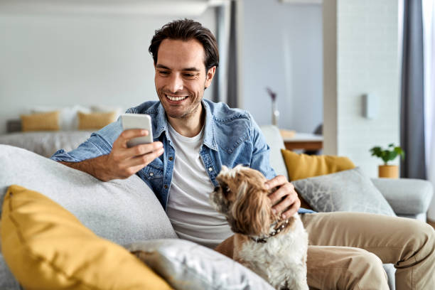 uomo felice che telefona al cellulare mentre si rilassa con il suo cane a casa. - men casual looking at camera one young man only foto e immagini stock